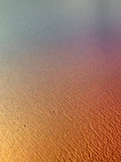 IMPACT 0017, extreme close-up of the paper's fine texture under a soft blue-grey to gold to pink colour wash, with a few tiny dark specks on the surface.