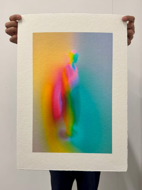 What Comes Back by Atelier Kalle Hellzén, hand-signed and numbered Limited Edition of 5, held in two hands against a pale wall to show its scale. The full sheet reads as an object, deckle edges loose at top and bottom, the figure rising through orange, pink and teal. Giclée on handmade kozo washi.
