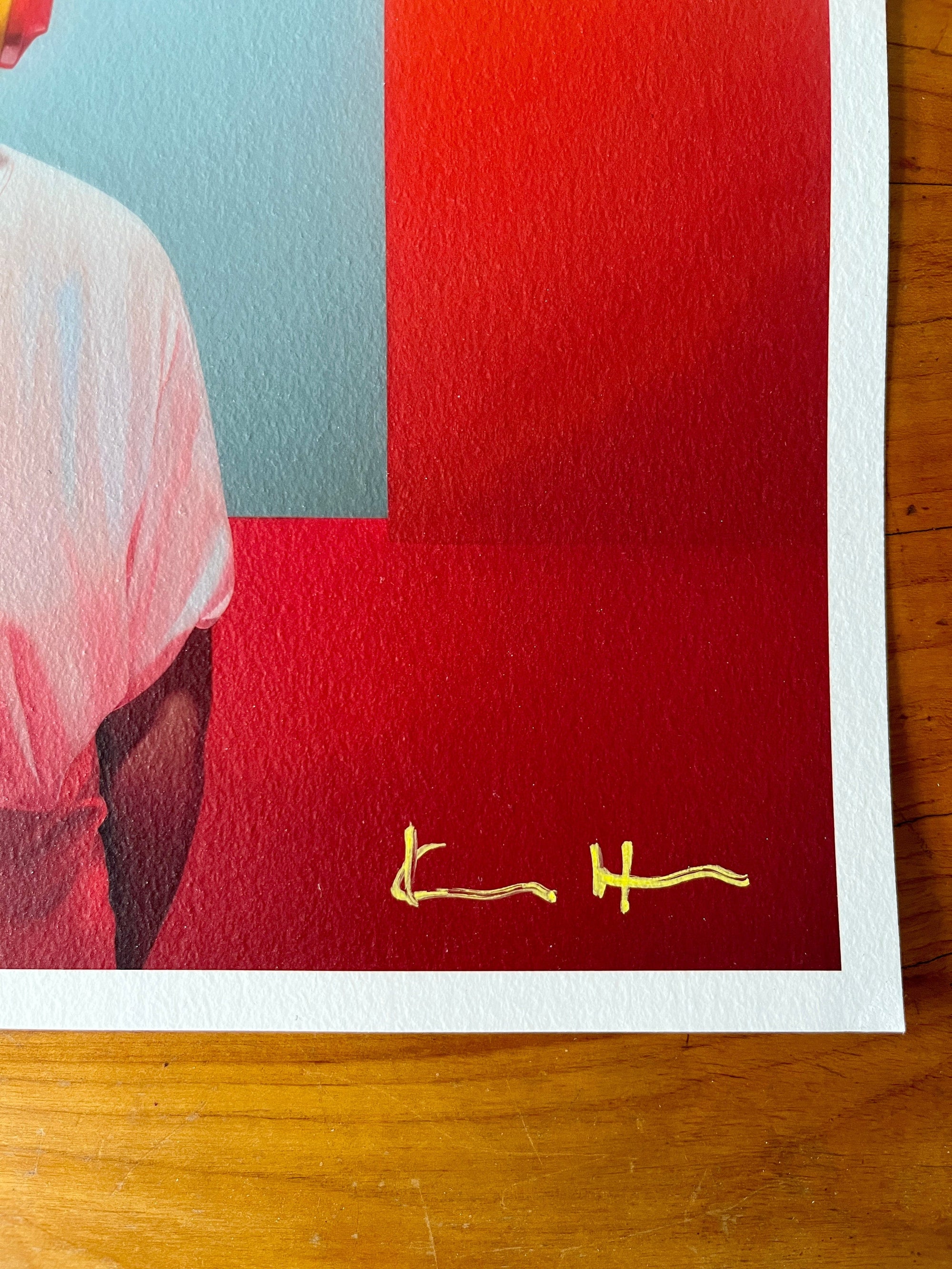 Lower-right corner detail of an unframed giclée print by Atelier Kalle Hellzén on a wooden tabletop, showing the hand-applied gold cursive signature.