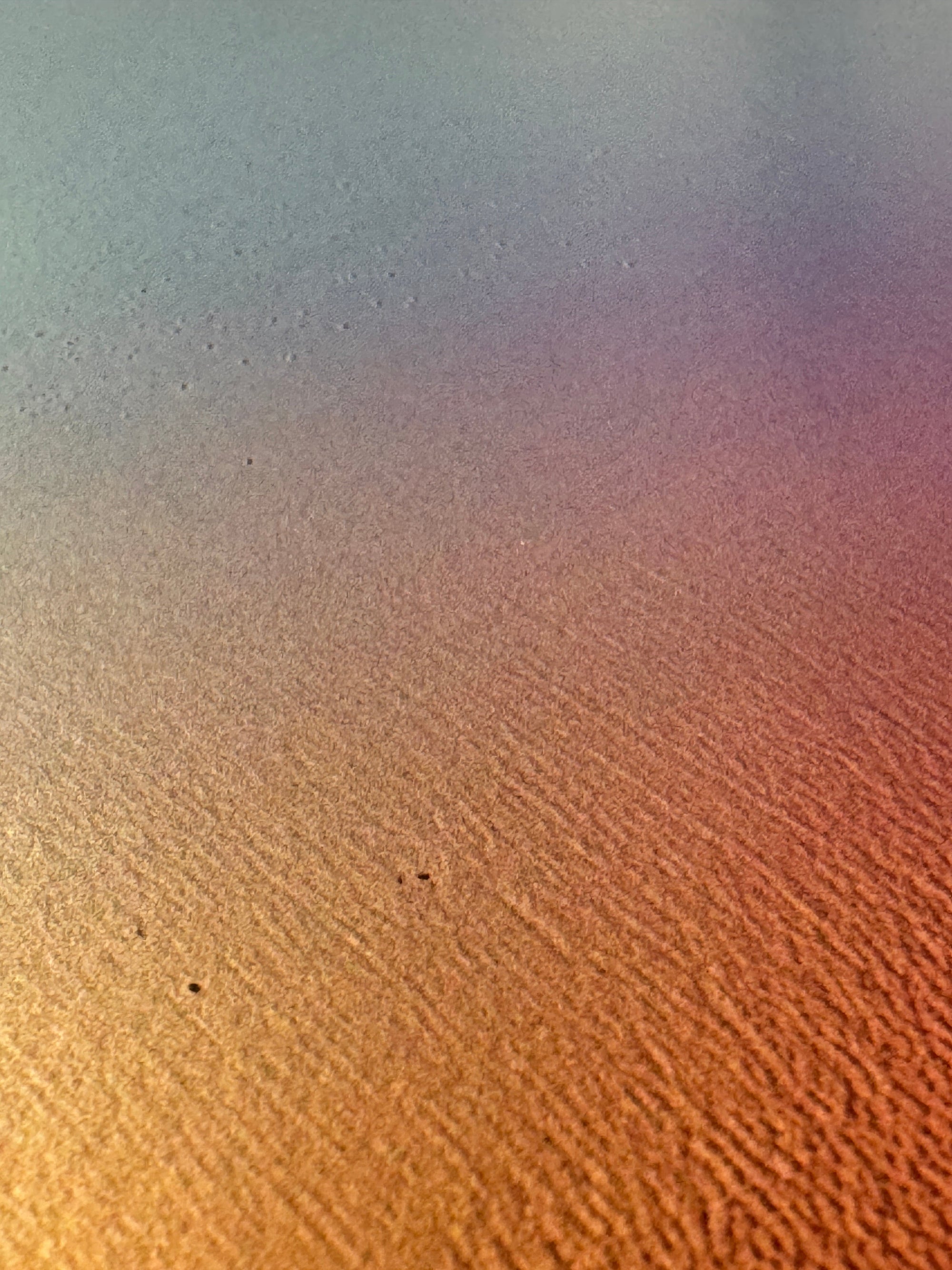 IMPACT 0017, extreme close-up of the paper's fine texture under a soft blue-grey to gold to pink colour wash, with a few tiny dark specks on the surface.