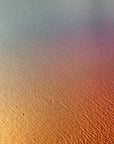IMPACT 0017, extreme close-up of the paper's fine texture under a soft blue-grey to gold to pink colour wash, with a few tiny dark specks on the surface.