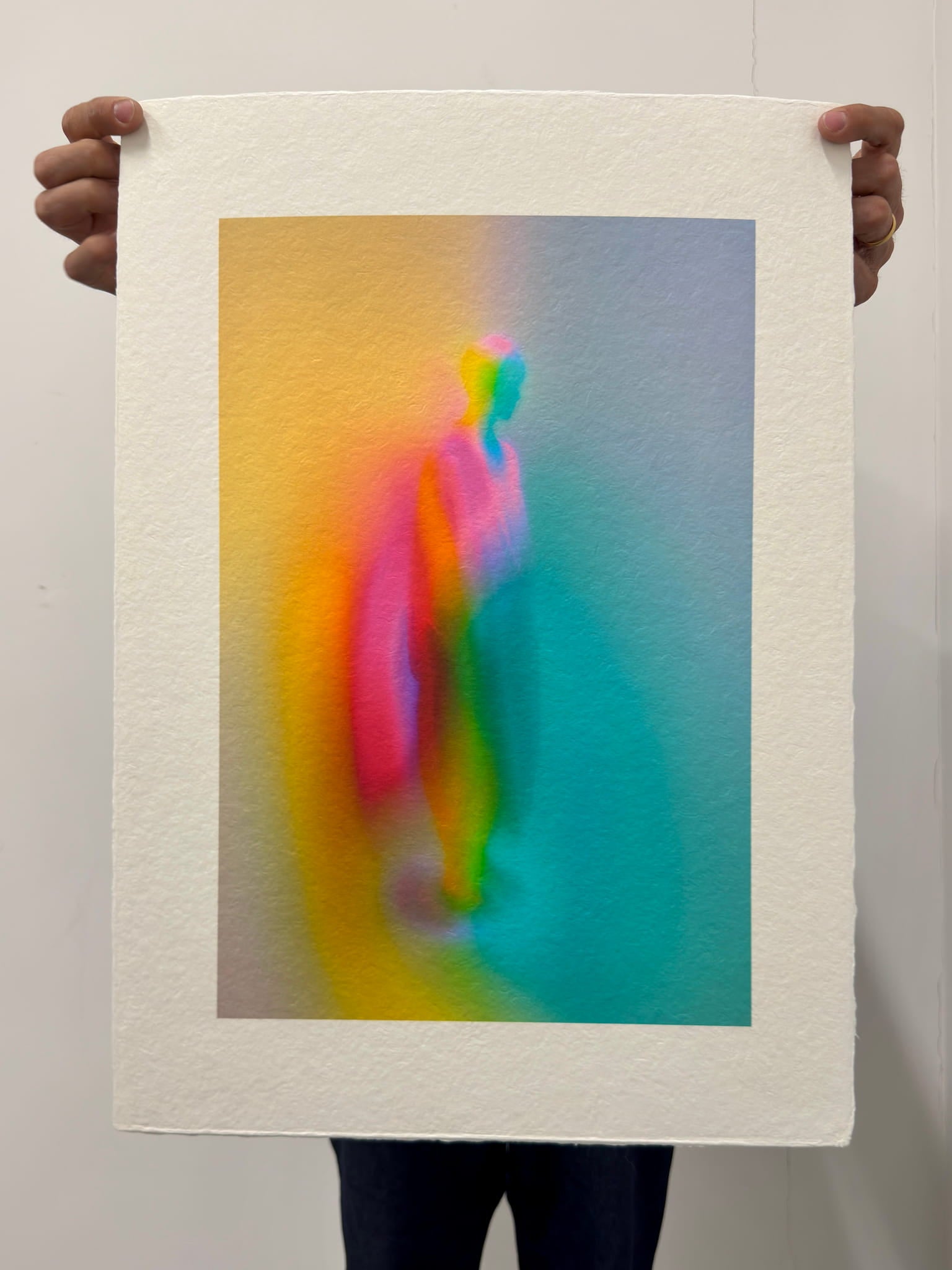 What Comes Back by Atelier Kalle Hellzén, hand-signed and numbered Limited Edition of 5, held in two hands against a pale wall to show its scale. The full sheet reads as an object, deckle edges loose at top and bottom, the figure rising through orange, pink and teal. Giclée on handmade kozo washi.