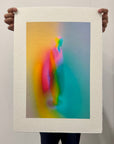 What Comes Back by Atelier Kalle Hellzén, hand-signed and numbered Limited Edition of 5, held in two hands against a pale wall to show its scale. The full sheet reads as an object, deckle edges loose at top and bottom, the figure rising through orange, pink and teal. Giclée on handmade kozo washi.
